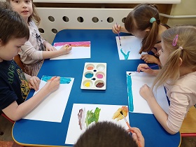 Dzieci siedzą przy stoliku i malują farbami po białej kartce papieru. 