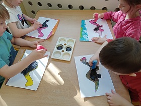 Dzieci siedzą przy stoliku i odciskają czarną farbę na kolorowych kartkach. Na kolorowych kartkach znajduję się szablon dinozaura. 