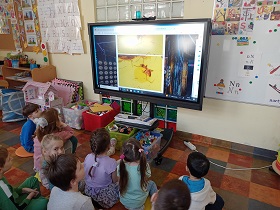Dzieci siedzą na podłodze na kolorowych poduszkach przed monitorem, na którym wyświetlane jest zdjęcie mrówki w bursztynie. 