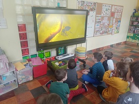 Dzieci siedzą na podłodze na kolorowych poduszkach przed monitorem, na którym jest przedstawiony film z mrówką w bursztynie. 