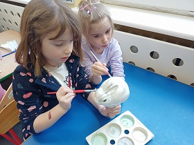 Dwie dziewczynki siedzą przy stole i malują farbami wspólnie białego, styropianowego królika.