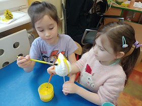 Dwie dziewczynki siedzą przy stole i malują żółtą farbą, wspólnie białą, styropianową kurę.