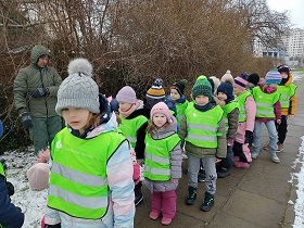 Dzieci ubrane w kurtki, czapki i zielone kamizelki stoją w parach na chodniku. Obok nich stoi pan w oliwkowej kurtce. 