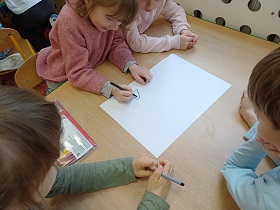 Troje dzieci siedzi przy stoliku. Dziewczynka w różowym swetrze rysuje czarnym flamastrem na białej kartce A3.