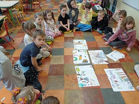 Dzieci siedzą w kółku na podłodze w sali. W środku kółka leżą ułożone kartki z rysunkami dzieci. Część dzieci się zgłasza. 