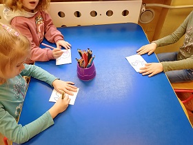 Trójka dzieci siedzi przy stoliku i koloruje kwiatki. Na środku stołu stoi pudełko z kredkami. 
