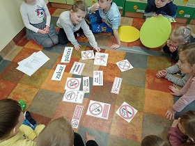 Dzieci siedzą w kółku na podłodze. NA środku kółka kółka znajdują się kartoniki mówiące o zakazie palenia. Jest też napis: Zakaz palenia. 