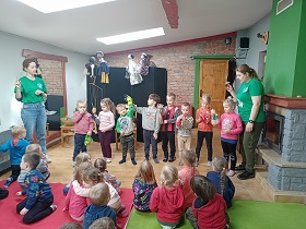 Dzieci siedzą na dywanach w sali i obserwują dwie panie w zielonych koszulkach oraz ósemkę dzieci z pacynkami na dłoniach 