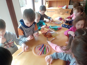 Dzieci stoją przy stole i ozdabiają drewniane łyżki kawałkami materiału, kolorowymi drucikami, cekinami.