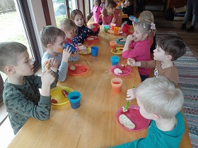 Przy stole siedzą dzieci i jedzą kiełbasę. Przed każdym dzieckiem ustawiony jest talerz i kubeczek.