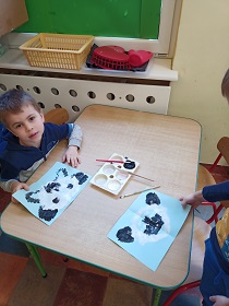 Przy stoliku siedzi chłopiec, który malował czarną i białą farbą, na niebieskiej kartce pandę. 