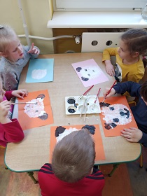 Przy stoliku siedzi piątka dzieci, która na kartkach maluje czarną i białą farbą pandy. 