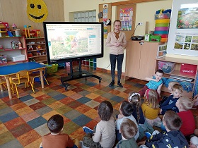 Na podłodze siedzą dzieci i spoglądają na nauczycielkę w beżowym swetrze, która stoi przy monitorze. Na monitorze wyświetlane jest koło z emocjami i obrazkami ukazującymi pogodę.