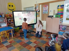 Na podłodze siedzą dzieci i spoglądają na chłopca w brązowej bluzce oraz na nauczycielkę w beżowym swetrze, która kuca przy monitorze. Na monitorze wyświetlane jest koło z emocjami i obrazkami ukazującymi pogodę.