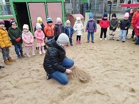 Dzieci stoją w kółku na placu zabaw, nauczycielka demonstruje wynik wsiąknięcia wody w piasek.