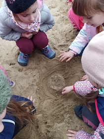 Cztery dziewczynki kucają na piasku i kopią w nim rękami. 