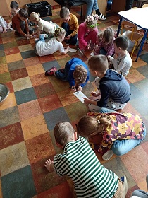 Dzieci w sali na podłodze, w grupkach rysują coś na kartkach.