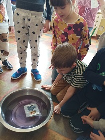 Na podłodze stoi metalowa miska wypełniona wodą. Wokół miski znajdują się dzieci obserwujące skutki położenia papierowego obrazka do wody. 