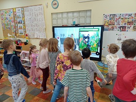 Dzieci stoją przed monitorem, na którym wyświetlany jest film z panią i kotem w trawie. 