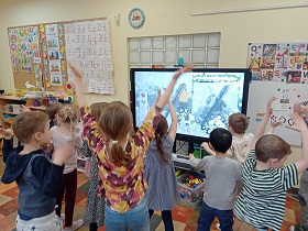 Dzieci wznoszą ręce do góry i patrzą na to, co jest wyświetlane na monitorze. 