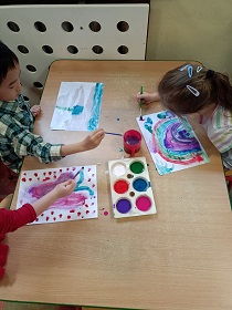 Stół, kolorowe farby, pędzle, chłopiec maluje niebo i niebieskiego kwiatka, dziewczynka maluje różowego motyla, druga dziewczynka maluje kolorowa tęczę
