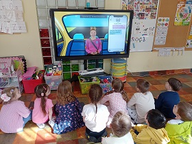 Dzieci siedzą przed monitorem w siadzie skrzyżnym i patrzą na monitor. Na nim wyświetlana jest pani w samochodzie oraz telefon i treść wiadomości. 