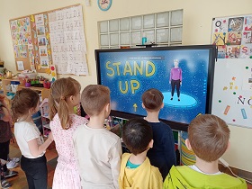 Dzieci stoją przed monitorem, na którym wyświetlany jest film z panią oraz napis stand up. 