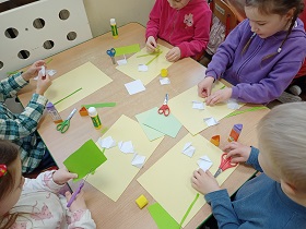 Piątka dzieci siedzi przy stoliku. Na nim znajdują się kartki - białe, zielone oraz blado żółte, nożyczki oraz kleje. Dzieci wycinają i przyklejają kwiatki do kartek. 