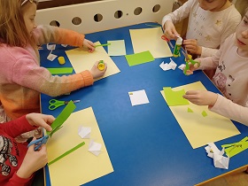 Czwórka dzieci siedzi przy stoliku. Na nim znajdują się kartki - białe, zielone oraz blado żółte, nożyczki oraz kleje. Dzieci wycinają i przyklejają kwiatki do kartek. 