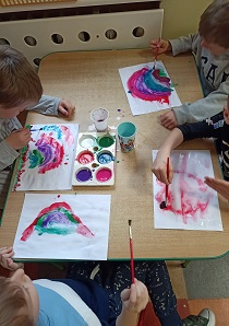 Stół, kolorowe farby, pędzle, 3 chłopców maluje kolorową tęczę, 1 chłopiec maluje serce