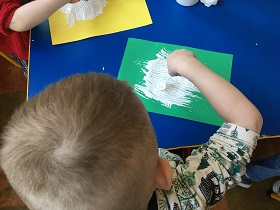 Chłopiec siedzi przy niebieskim stoliku i maluje białą farbą po zielonej kartce.