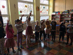Dzieci stoją w półkolu i podnoszą jedną rękę do góry.