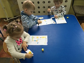 trójka dzieci siedzi przy stoliku i przykleja odpowiednie obrazki na kartę pracy.
