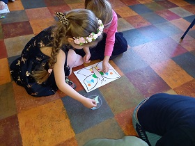 Na podłodze siedzą dwie dziewczynki. Dziewczynka po lewej stronie wyjmuje z pojemnika zielone półkole, zaś dziewczynka po prawej stronie kładzie na skrzydle motyla zielony trójkąt.