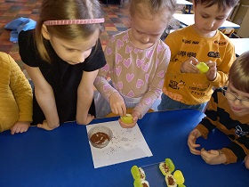 Na stoliku położona jest serwetka z ustawionym pojemniczkiem z nasionami. Dziewczynka trzyma w jednej dłoni żółty pojemnik a drugą sypie ziarenka. Obok stoją dzieci i się przyglądają.