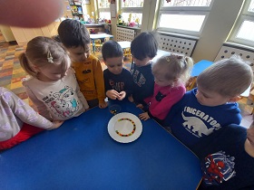 Dzieci stoją wokół stolika na którym ustawiony jest talerz z ułożonymi kolorowymi cukierkami. Chłopiec w niebieskiej bluzce trzyma w dłoni cukierek.