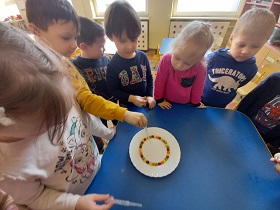 Dzieci stoją wokół stolika. Chłopiec w żółtej bluzie trzyma w dłoni pipetę, która wlewa wodę na talerz.