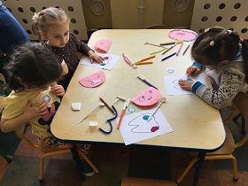 Dzieci wykonują pracę plastyczną przy stoliku. Na stoliku leżą różowe kartki w kształcie półkola, flamastry oraz lusterka.