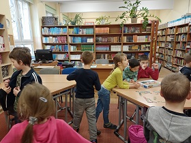 Dzieci stoją i siedzą w bibliotece szkolnej. Za dziećmi znajdują się regały z książkami. 