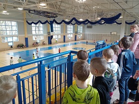 Dzieci stoją na trybunie i obserwują dzieci, które biegają na sali gimnastycznej i grają w piłkę nożną.