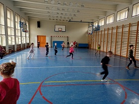 Dzieci biegają po sali gimnastycznej i grają w piłki. Sala gimnastyczna ma niebieską podłogę i namalowane linie. Znajduję się w niej również bramka i kosze go grania w koszykówkę. 
