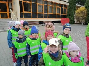 Dzieci w kurtkach, czapkach i zielonych, odblaskowych kamizelkach stoją przed budynkiem, ustawione w parach. 