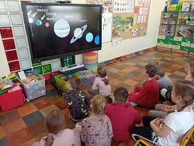 Dzieci siedzą na podłodze i krzesełkach przed monitorem. Na monitorze wyświetlany jest film, na którym widać planety . Napisy: Wenus, Ziemia, Mars, Jowisz, Saturn. 