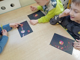 Trzech chłopców siedzi przy stoliku i wydrapuje żółtymi patyczkami układ słoneczny na czarnych kartkach.