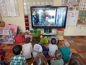Dzieci siedzą na podłodze przed monitorem, na którym wyświetlany jest film, w którym kobieta ma uniesione do góry wszystkie włosy i znajduję się w statku kosmicznym. 