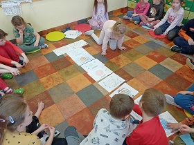 Na podłodze siedzą dzieci w kółeczku, na kolorowych poduszkach. W środku kółka znajdują się ułożone w pionie rysunki planet. Jedna dziewczynka znajduje się blisko ilustracji. 