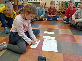 Dziewczynka w fioletowej bluzce i spódniczce siedzi na podłodze i piszę kredką coś na kartce. Przed nią leżą kartki ułożone w pionowym rzędzie. Za dziewczynką siedzą chłopcy i na nią spoglądają. 