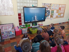 Dzieci siedzą na podłodze i krzesełkach przed monitorem. Na monitorze wyświetlany jest film, na którym widać start rakiety kosmicznej. 