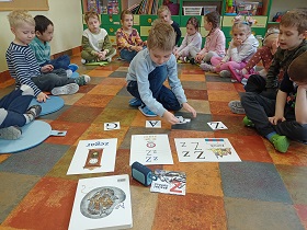 Chłopiec siedzi i układa kartoniki z literkami. Przed nim leżą ilustracje z literką z, głośnik oraz książka. Za chłopcem w półkolu siedzą pozostałe dzieci. 