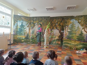 Pan i pani w strojach teatralnych stoją na tle dekoracji leśnej. Dzieci spoglądają na nich siedząc na widowni. 